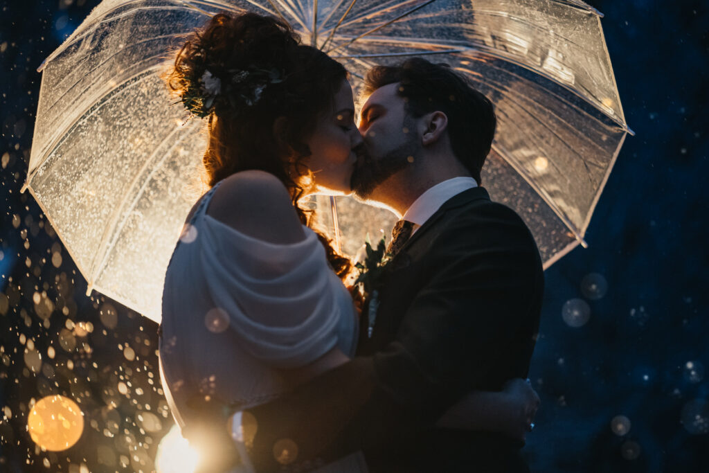 Braut und Bräutigam küssen sich unter dem Regenschirm, es ist dunkel und es regnet.