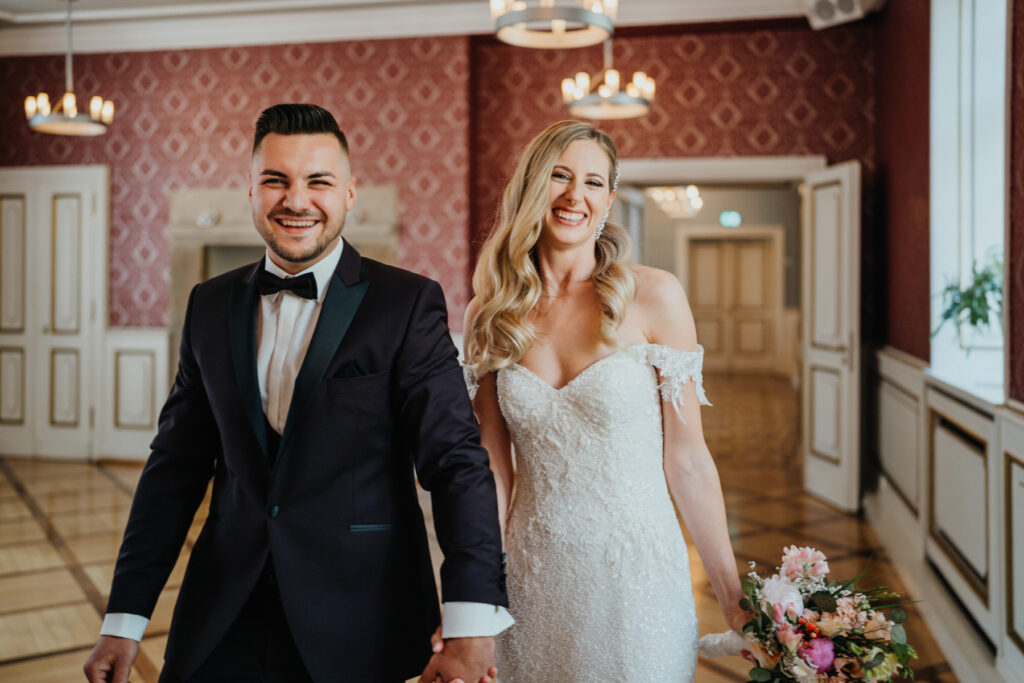 Die Braut und der Bräutigam gehen Hand in Hand aus dem Standesamt raus nach der Trauung und lächeln voller Freuede.