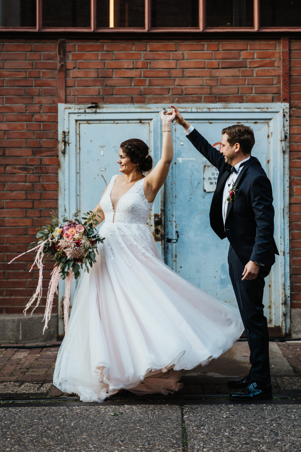 Die Braut tanzt mit dem Bräutigam hinter einer blauen industriellen Tür und hält den Brautstrauß in der Hand
