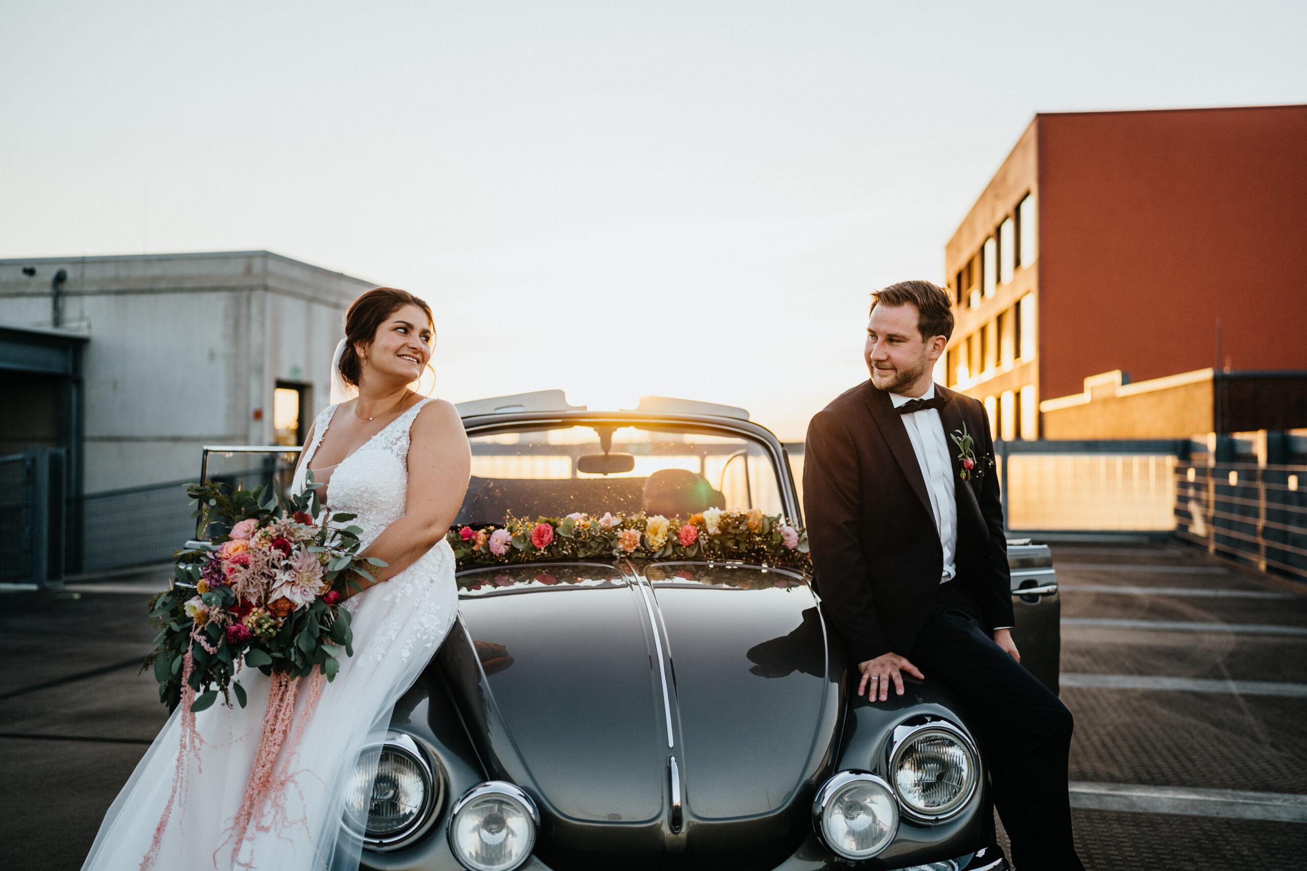 Das Brautpaar lehnt sich am Auto an und lächelt sich an, die Braut hält den Brautstrauß